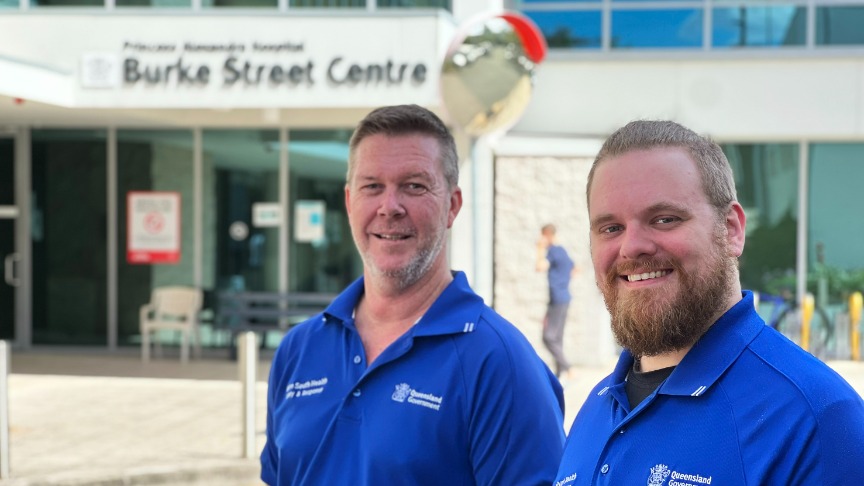 Two male security officers wearing blue shirts provide customer service to outpatients outside PA Hospital’s Burke Street Centre entrance.