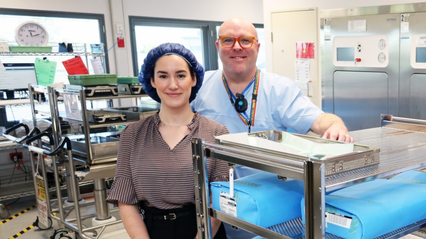A female clinician wearing a hairnet and a male nurse wearing scrubs stand in a sterile cleaning area alongside racks of blue wrapped instrument trays