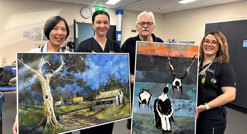 Four healthcare workers hold two paintings in a hospital room. One painting depicts a rural scene with a tree and cabin, the other shows two birds.