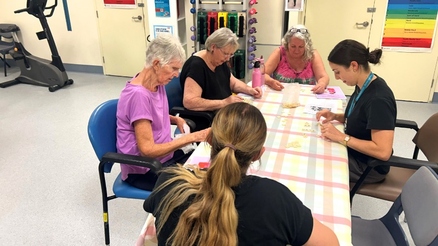 A group of older women sitting at a table in a gym, engaged in a group program activity