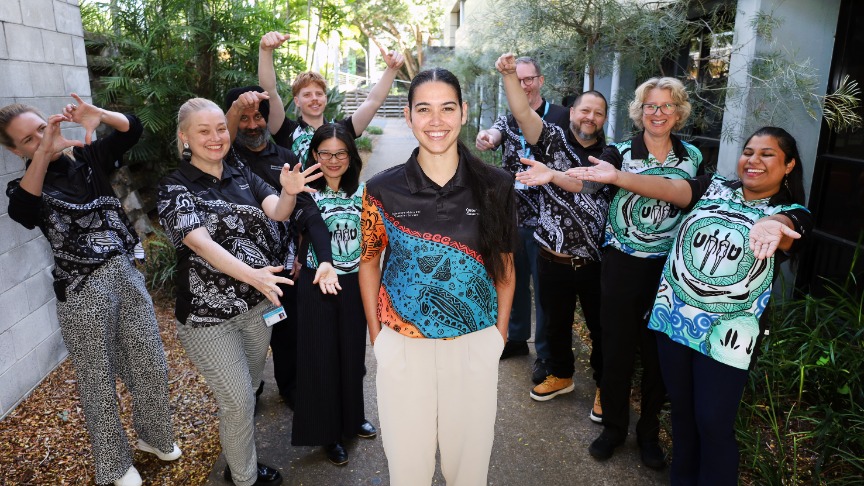 An Aboriginal woman stands in front of a multicultural team of clinicians wearing identifiable shirts outside a hospital building who celebrate her with hand gestures