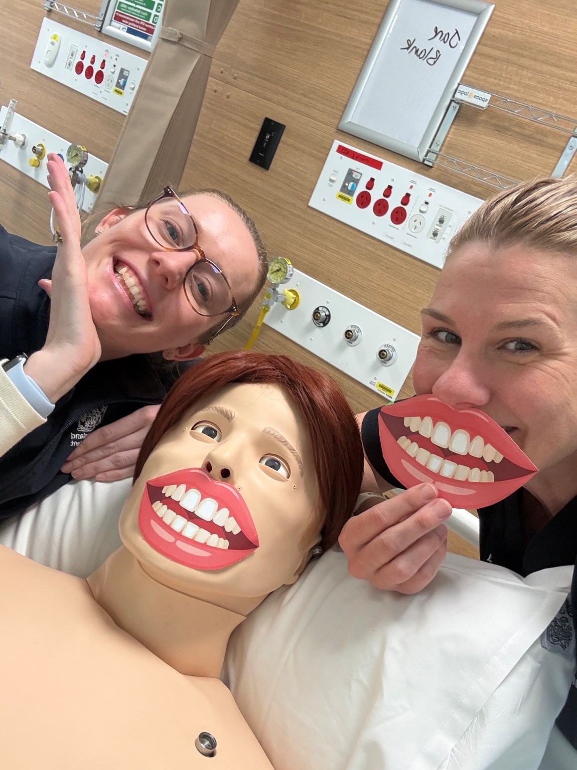two nurses on either side of a simulation mannikin. One nurse and mannikin are holding 'smile' props