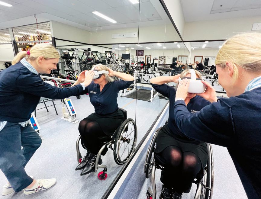 A clinician positions a virtual reality headset on a woman in a wheelchair with the action reflected in a mirror