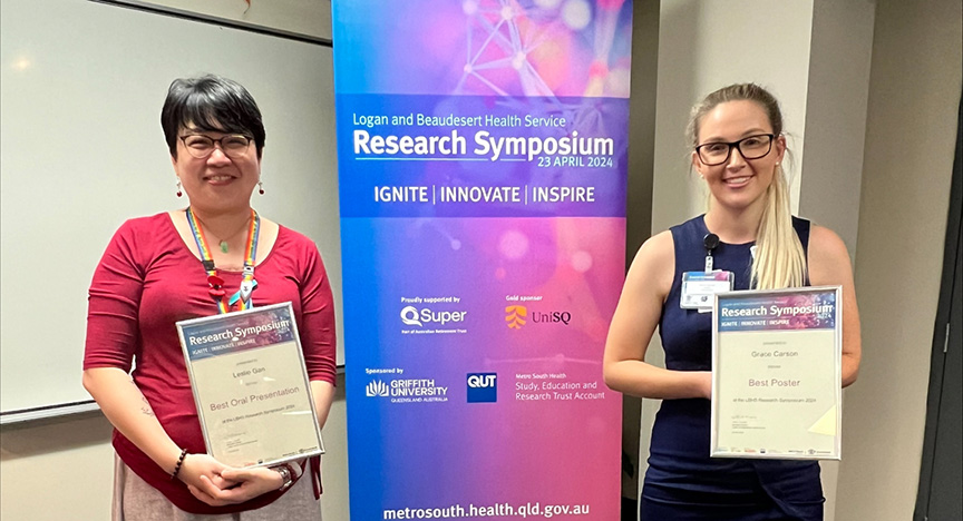 Two individuals holding certificates stand in front of a banner for the Logan and Beaudesert Health Service Research Symposium on April 23, 2024.