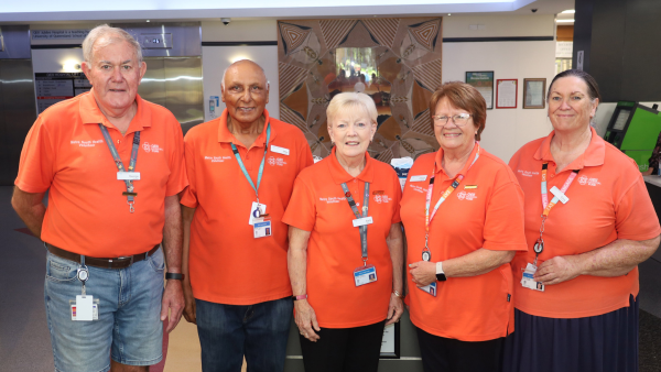 Five volunteers in orange shirts stand together in a hospital lobby, wearing name tags and lanyards.