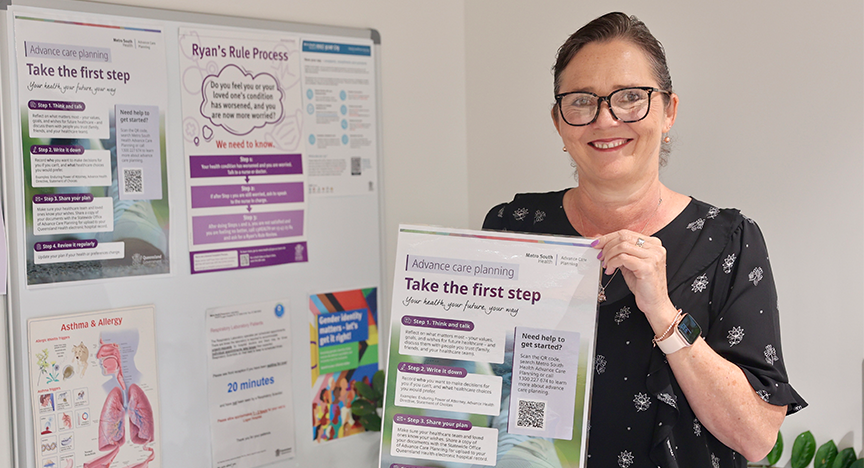 A smiling woman with glasses holding up a poster standing in front of a notice board