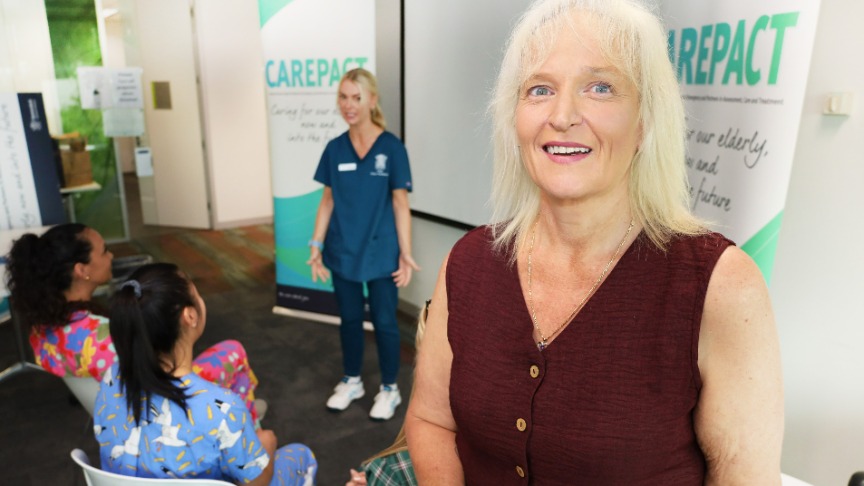 A female nurse practitioner smiles at the camera at a masterclass event hosted in a training room attended by other female nurse practitioners