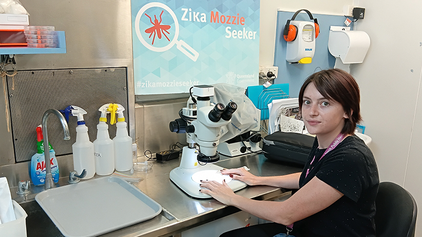 A young woman with shoulder-length brown hair, sitting in a lab in front of a microscope and looking at the camera. There is a large blue sign above that reads Zika Mozzie Seeker with a picture of a mosquito inside a magnifying glass.
