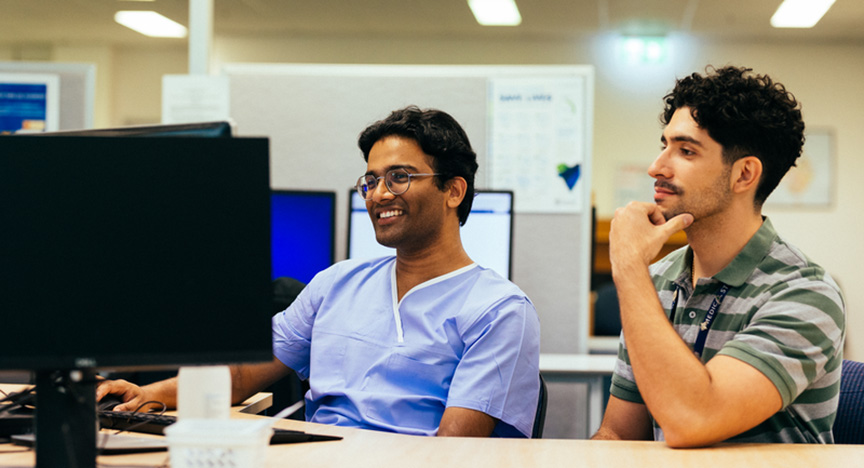 Two people are sitting at a desk in an office, looking at computer screens. One is wearing a blue scrub top, and the other is in a striped shirt.