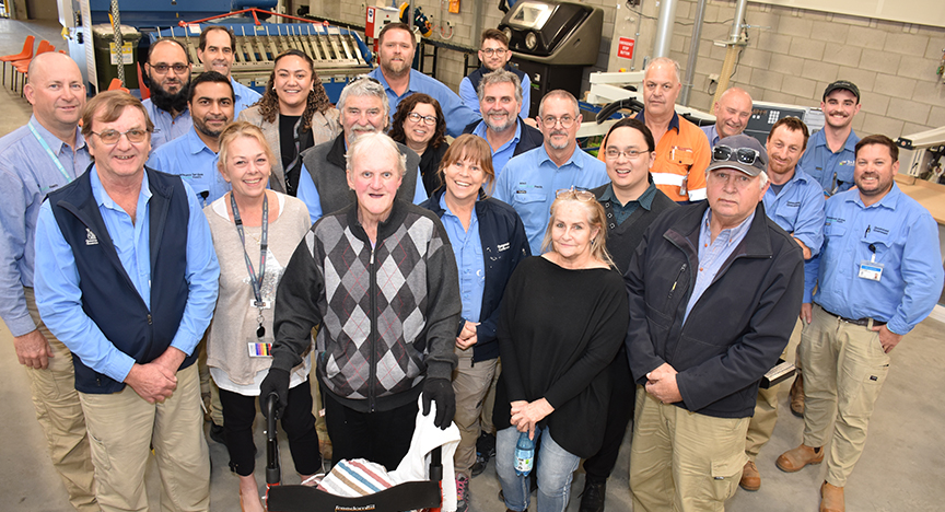 A group of people in work attire standing together in a workshop, with machinery and tools in the background.