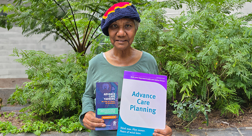 An elderly First Nations woman smiling and holding an advance care planning document and an advance care yarning brochure outside in front of greenery