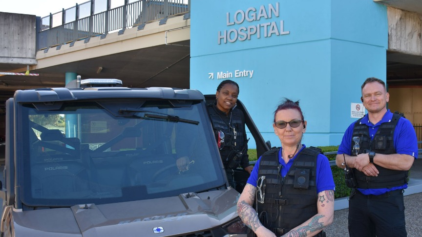 Three security guards wearing blue polo shirts and new black vests stand in front of a buggy and Logan Hospital sign