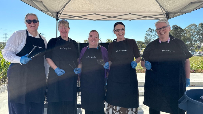 Logan and Beaudesert Executive staff holding up BBQ utensils, standing in front of a BBQ