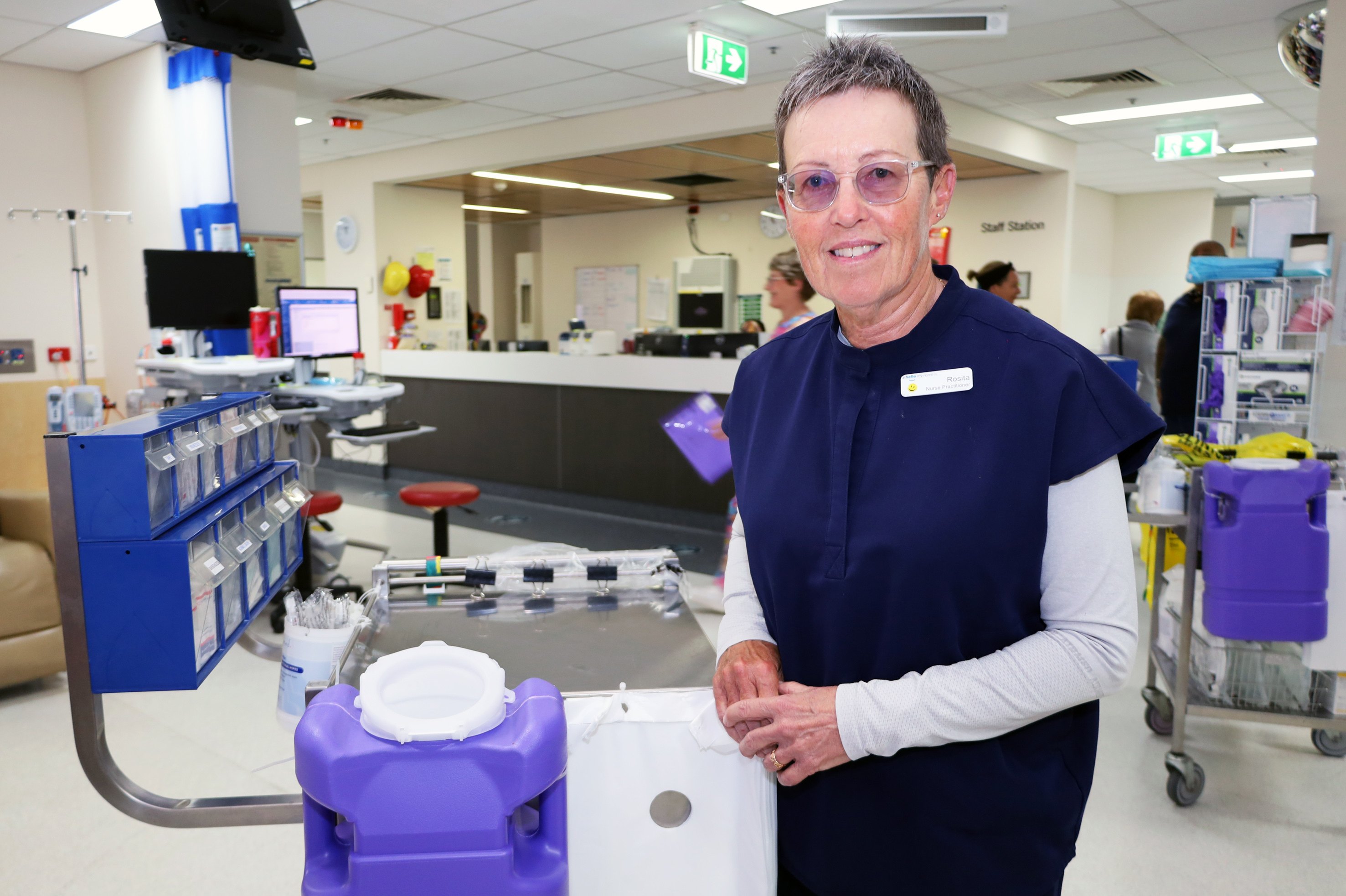An experienced female nurse wearing scrubs standing in a clinic surrounded by equipment used to deliver cancer therapy