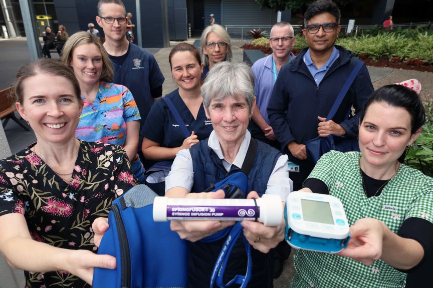 a group of nine clinicians in an outdoor space wearing scrubs and uniforms reaching up to the camera holding nursing items used in the treatment of patients in the community