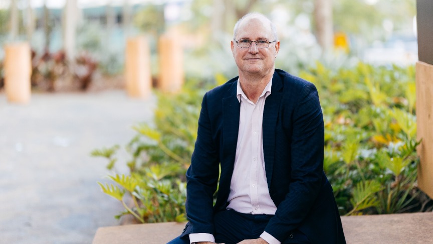 A man in a suit and pink shirt sits on a bench, looking thoughtfully into the distance