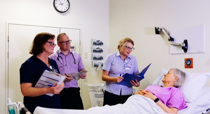 Three healthcare professionals are discussing a patient's condition at the bedside in a hospital room. The patient is lying in bed.