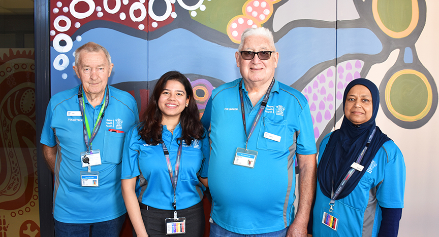 Four volunteers in blue shirts stand in front of a colorful mural at Eight Mile Plains Satellite Hospital.