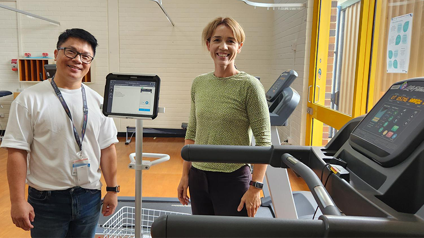 A man and a woman standing inside a gym next to a treadmill with an iPad displayed between them.
