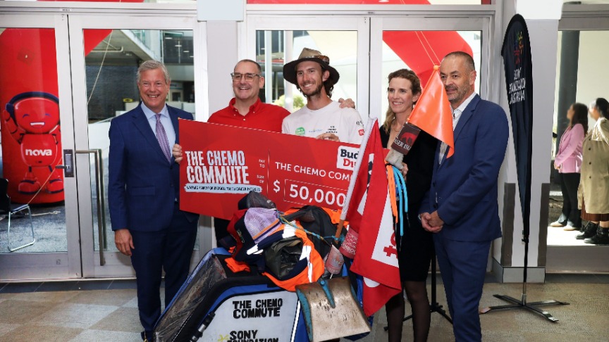 Marcus, a former cancer patient, stands with supporters in front of a large cheque and a sign celebrating his chemo commute