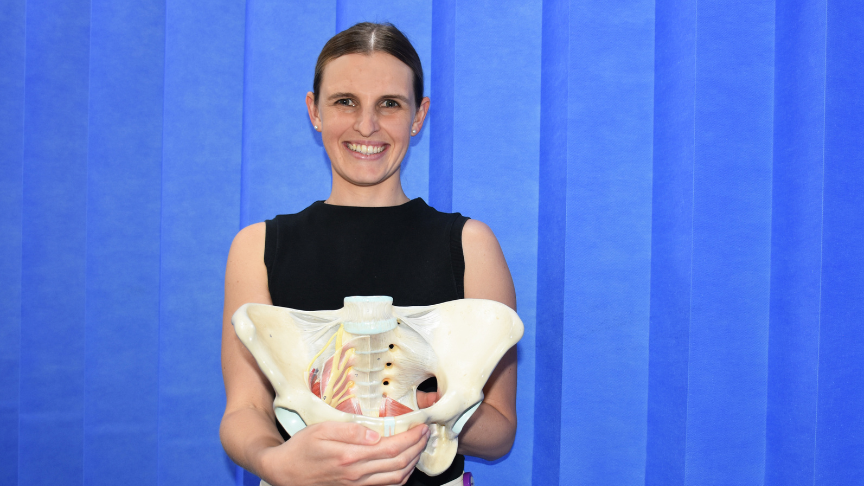 Picture of a female physio holding a pelvis teaching aide in front of a blue curtain
