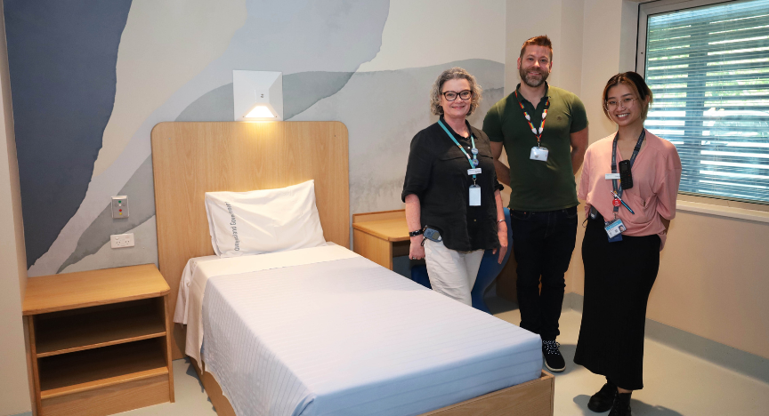 Three healthcare workers stand in a newly refurbished hospital room with a single bed, bedside table, and window.