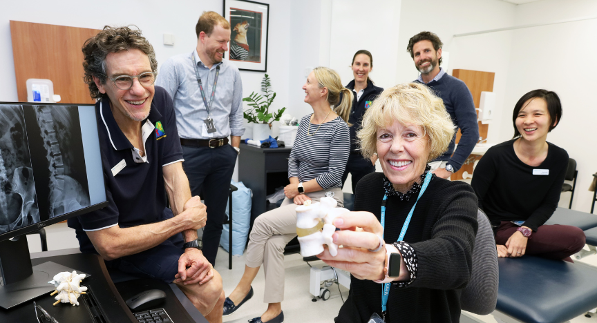 A group of physiotherapists in a treatment room with an experienced female physiotherapist at front of shot holding a model of vertebra. A male physiotherapist leans on the edge of desk with CT image of a spine on the computer. Two male and three female physios interact with each other in the background.