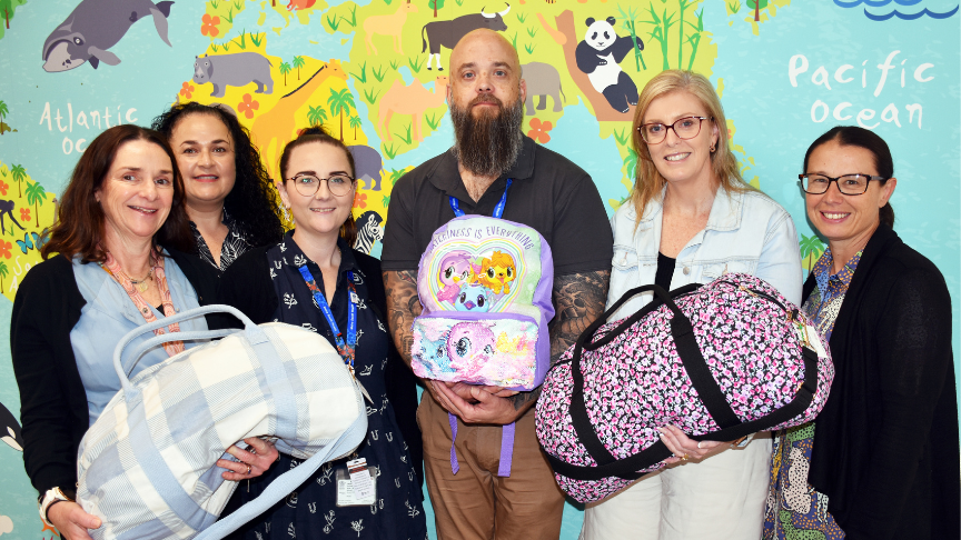 Several individuals, some holding overnight bags standing in front of a wall with a bright world map cartoon mural on it