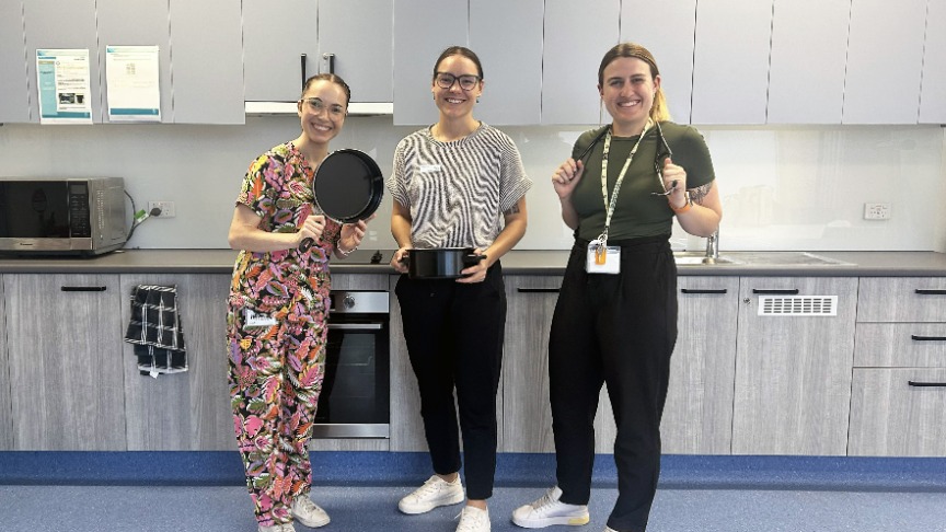 Three women in a kitchen, holding a pan, engaged in cooking together as part of the Older Adults Liaison team