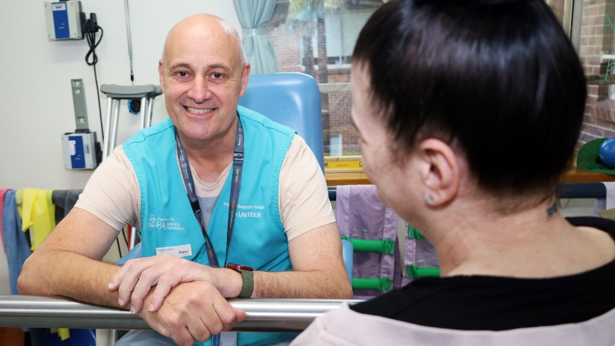 A male volunteer wearing an identifying vest interacts with a female patient in a hospital rehabilitation gym