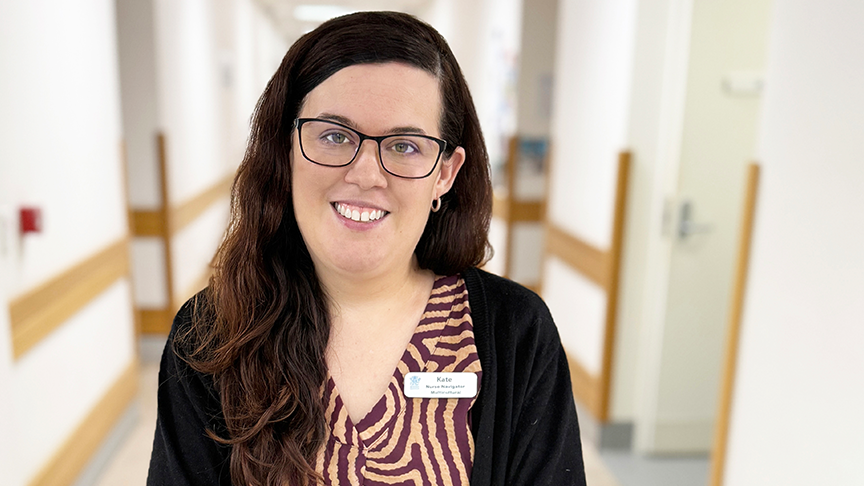 A woman with brown wavy hair, standing in the middle of a hospital corridor. She is wearing glasses, a patterned top and cardigan and has a smile on her face.