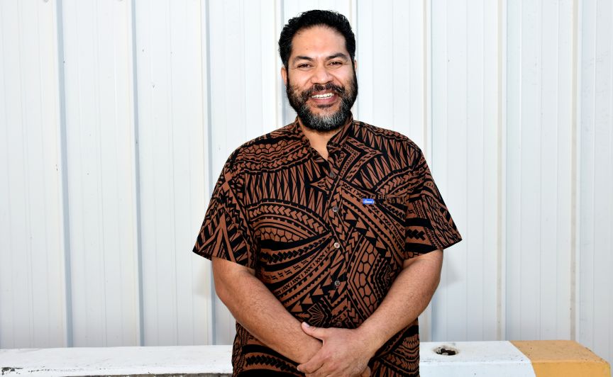 Samoan Man in a black and brown shirt, standing in front of an external white wall 