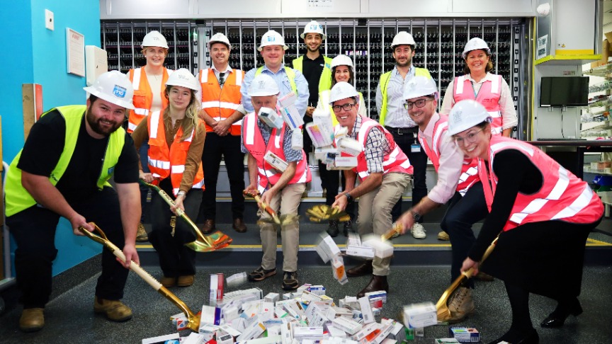 Hospital staff in hard hats and vests participate in pharmacy expansion, holding golden shovels near a pile of boxes