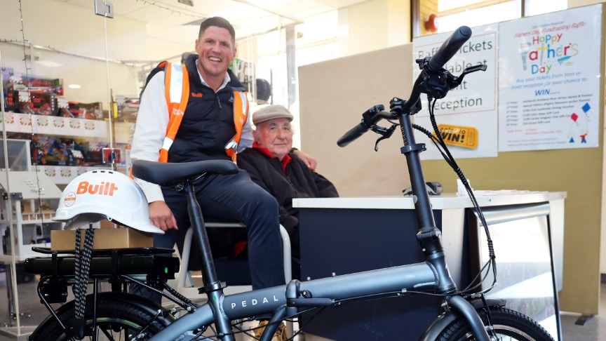 two men sit behind a staged black e-bike.