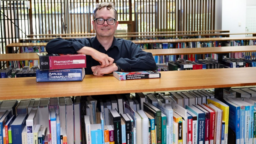 A man with glasses, Pharmacy Educator Karl Winckel, stands in the library surrounded by books