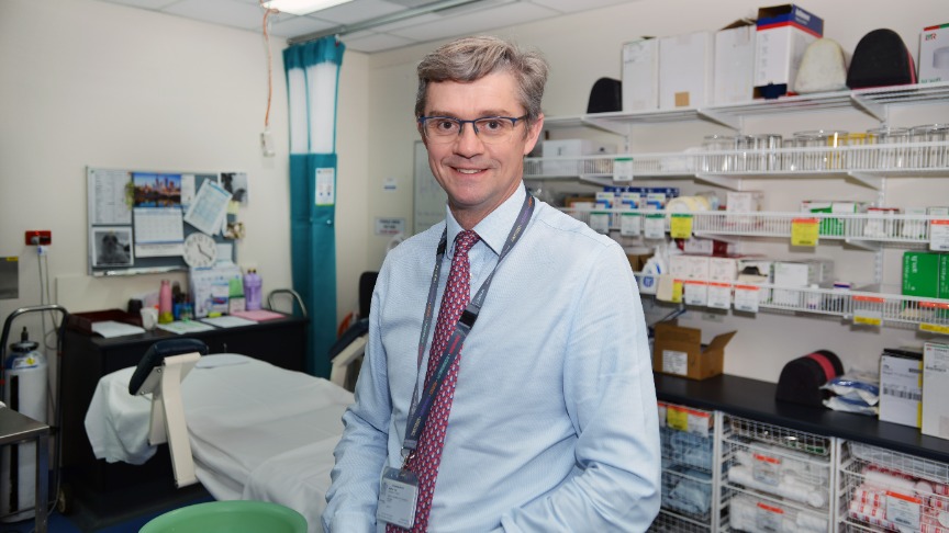 An experienced male doctor smiling at the camera with a backdrop of an orthopaedics clinic procedure room