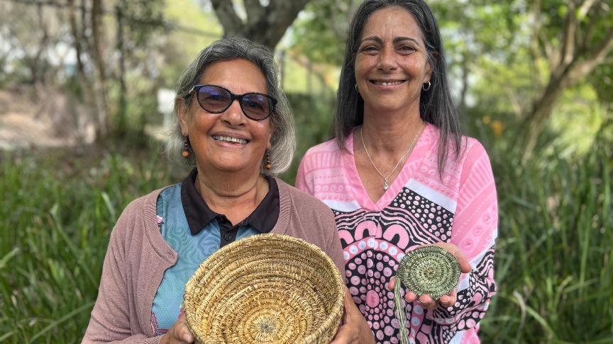 Two women demonstrate basket weaving at a NAIDOC Week event, surrounded by trees and showcasing their crafted baskets