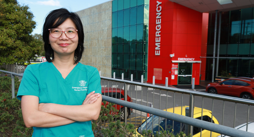 A nurse practitioner in teal scrubs stands with arms crossed outside an emergency department entrance.
