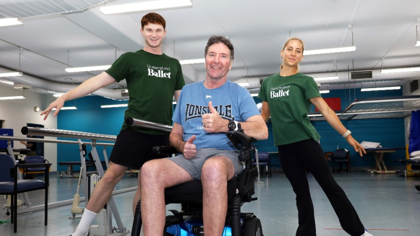 A man in a wheelchair at a hospital, accompanied by two ballet instructors in the background