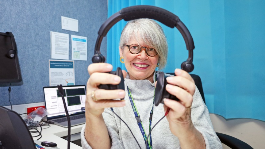 An experienced female allied health professional with headphones is working at a desk in a hospital clinic.