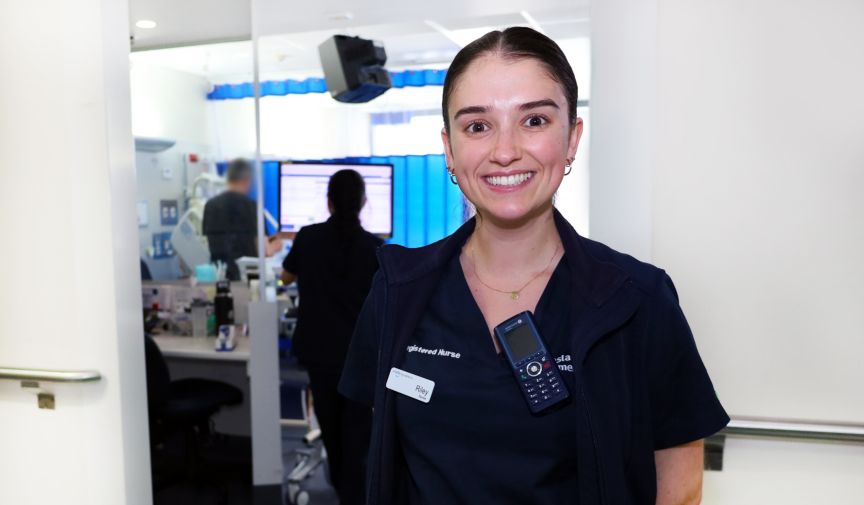 A female nurse in uniform standing in a high dependency ward with other nurses in the background working on bedside computer 