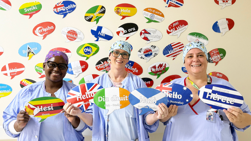 Three women wearing operating theatre PPE holding out laminated speech bubbles in front of a wall