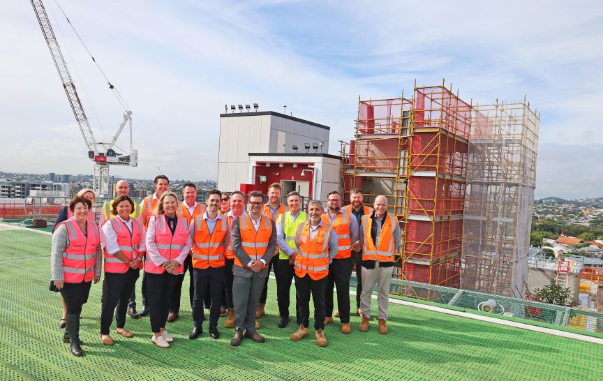 A group of 17 men and women wearing high visibility vests standing on a helipad in front of construction scaffolding and cranes with a suburban view 