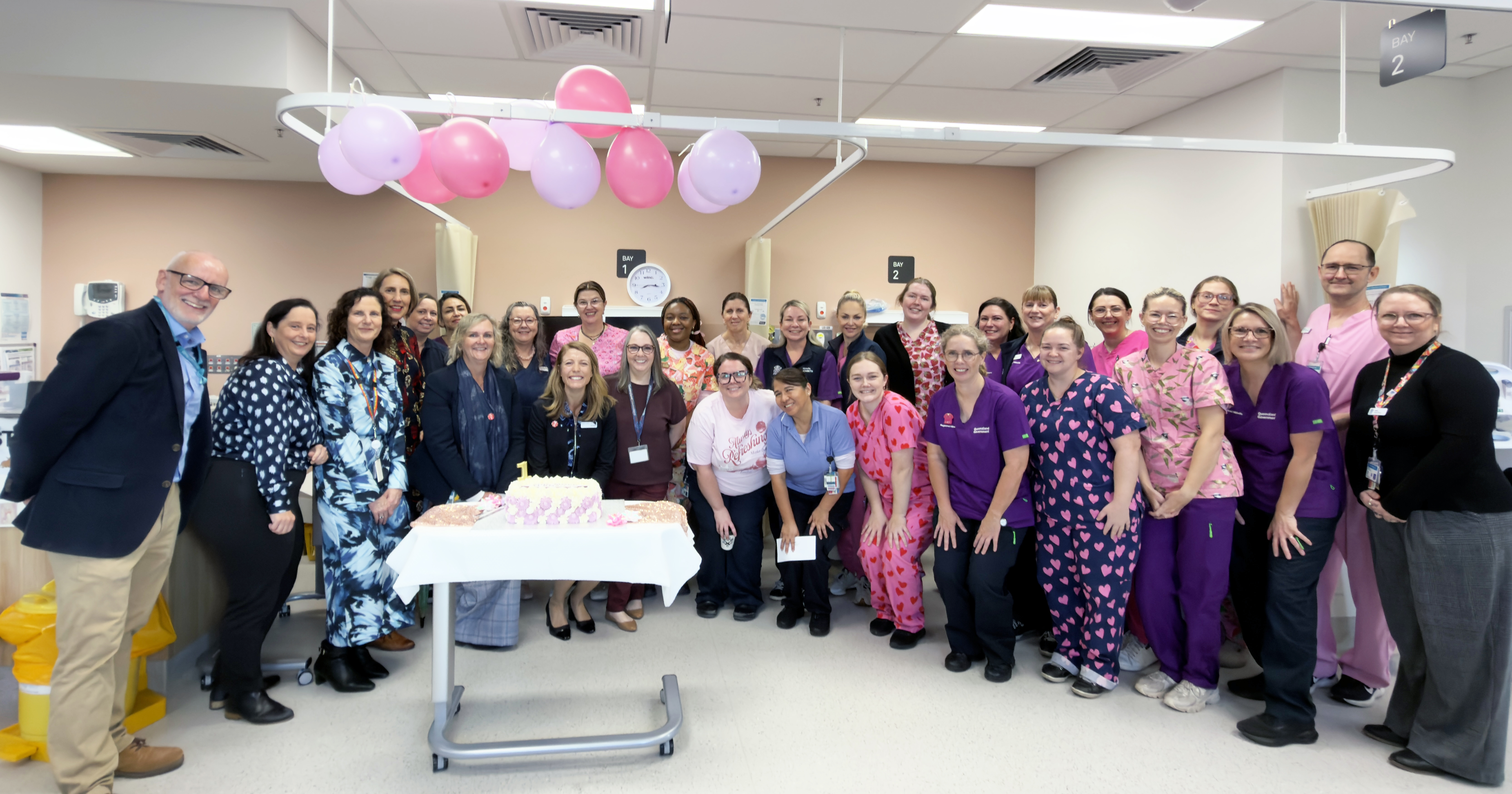 Large group of nusring staff and executives standing in a clinical area, behind a birthday cake