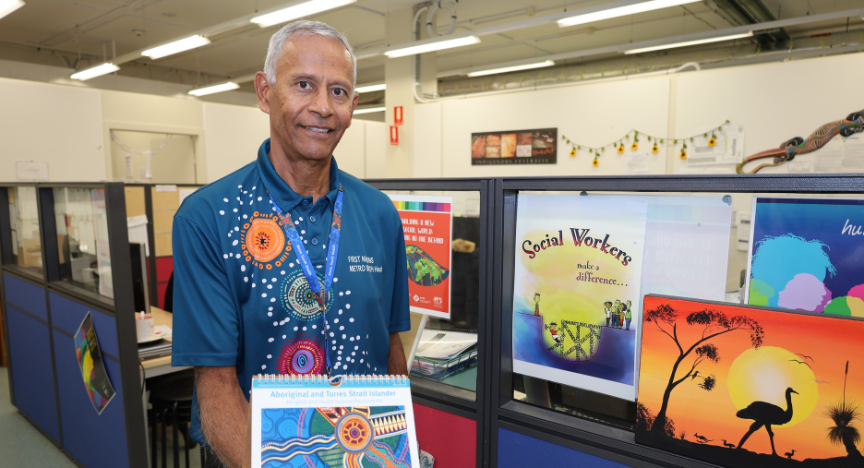 A person in a decorated shirt holds a First Nations Health booklet in an office with colorful posters about social work and First Nations culture.