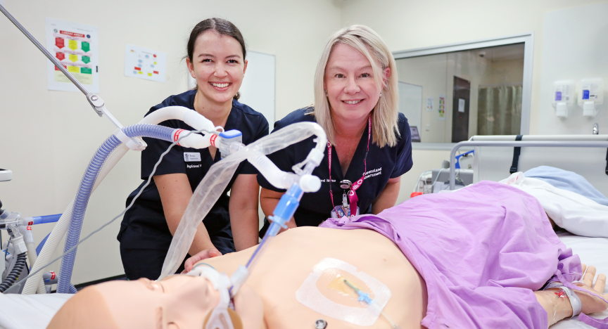 Two women in scrubs, Libby and Emma, pose next to a medical mannequin in a healthcare environment