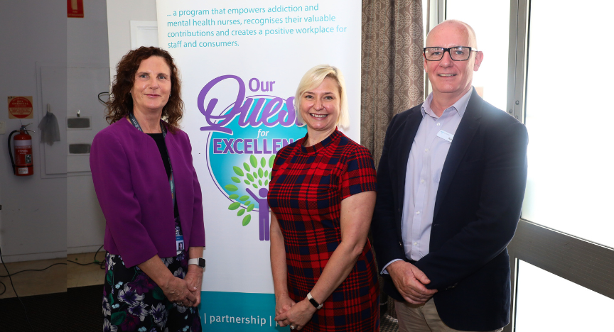 Three people stand beside a banner reading Our Quest for Excellence, smiling at the International Nurses Day event